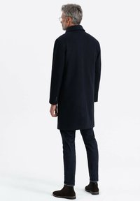 Cappotto di lana blu navy con design a un solo petto, colletto a discontinuità, tasche laterali e una texture liscia, indossato sopra jeans scuri e stivali.