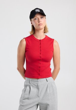 Jonge vrouw die een rood mouwloos gebuttoned topje draagt, lichtgrijze broek en een zwarte baseballpet staand tegen een effen lichte achtergrond.