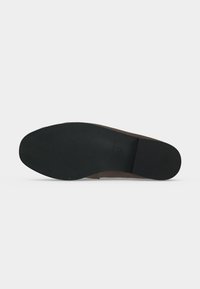 Semelle d'une chaussure habillée en cuir marron avec grip en caoutchouc noir et petit texte embossé sur la zone de la voûte plantaire sur un fond gris uni.