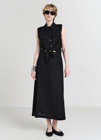 Maglietta nera senza maniche con due tasche sul petto e cintura annodata in vita, abbinata a una lunga gonna nera e scarpe flatte nere. Bracciale argento in evidenza.