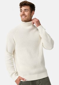 Pull à col roulé couleur crème avec un motif de tricot texturé, des poignets et un ourlet côtelés. Le modèle le porte dans une pose décontractée, la main sur le col.