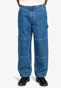 Pantaloni in denim blu a gamba larga con dettagli a pannello cuciti e tasche laterali. Presentano una vita alta e una vestibilità ampia, realizzati in robusto tessuto di cotone.
