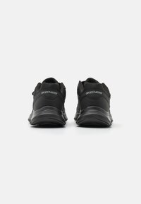 Crne sportske cipele s teksturiranim mrežastim gornjim dijelom, podstavljenom potplatom i podignutim petom. Imaju suptilno "SKECHERS" brendiranje na peti.