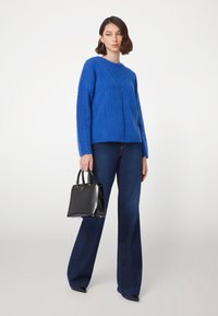 Maglione lavorato a maglia blu royal con motivo a treccia, abbinato a jeans svasati blu scuro. La modella tiene una borsa a tracolla in pelle lucida nera.