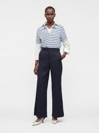 Femme portant un pull à manches courtes rayé bleu sur une chemise blanche, un pantalon large à rayures bleu marine et des chaussures blanches à talon bas.
