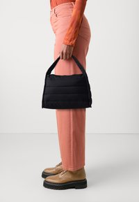 Sac à main matelassé noir avec une forme structurée, doté d'une petite poignée. Porté avec un pantalon large rose et des chaussures à lacets beiges.