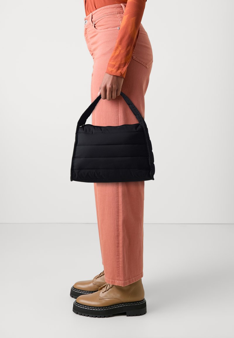 Sac à main matelassé noir avec une forme structurée, doté d'une petite poignée. Porté avec un pantalon large rose et des chaussures à lacets beiges.