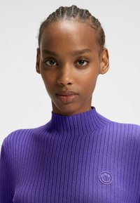 Lila gerippter Rollkragenpullover mit enganliegendem Design. Verfügt über ein kleines rundes Logo auf der linken Brust. Glatte Textur.