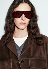 Persona con lunghi capelli scuri che indossa grandi occhiali da sole tartaruga, una giacca in suede marrone, un maglione grigio e una camicia bianca contro uno sfondo neutro.