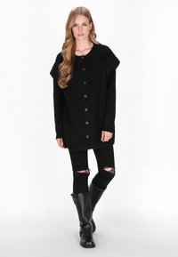 Czarny oversize sweter z teksturowanej tkaniny, z dużymi guzikami z przodu i otwartymi rękawami. W zestawie z czarnymi przetartymi rurkami i wysokimi botkami.