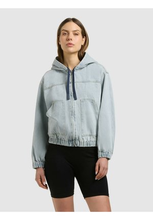 Moteris dėvi šviesiai mėlyną džinsinę su užtrauktuku ir tamsiomis raištelėmis, juodus dviračių šortus, stovinti priešais paprastą foną.