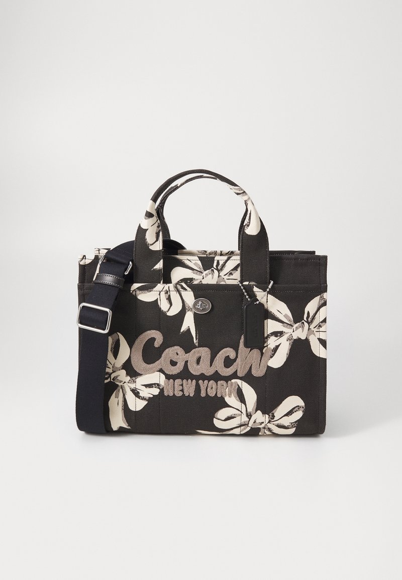 Sac cabas floral noir et blanc Coach avec double anses, logo devant, bandoulière et fermeture à tourniquet sur un fond uni.