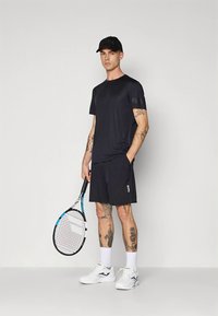 Juoda sportinė marškinėliai ir šortai, abu su logotipu. Žaidėjas laiko teniso raketę, dėvi baltus batus ir juodą kepurę.