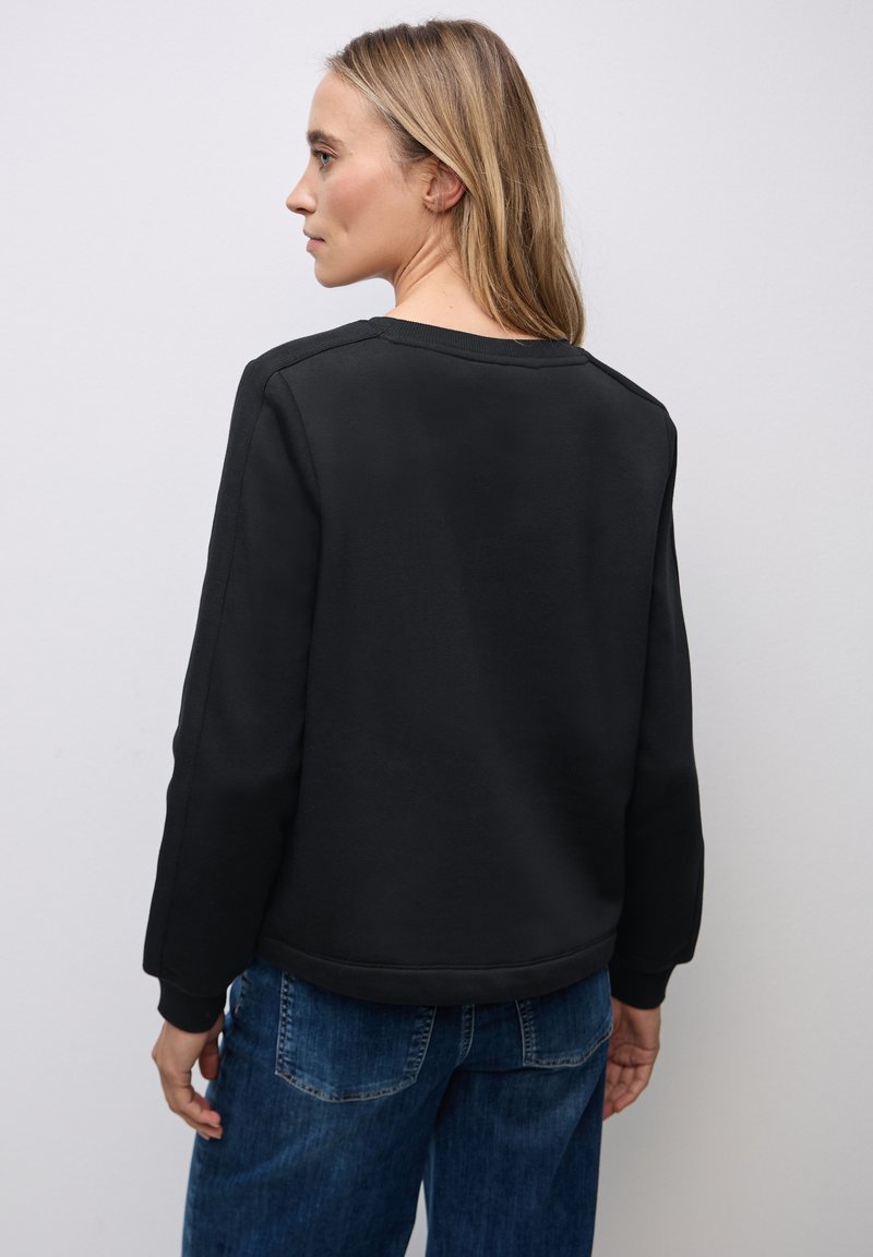 Felpa nera con collo a giro e maniche lunghe, caratterizzata da una texture liscia. Indossata con jeans blu casual, mostrando una vestibilità rilassata.