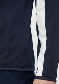 Polo de culoare bleumarin cu mânecă lungă, având o dublă fâșie albă în diagonala pe braț, realizat dintr-un material neted, elastic și cu un design ajustat.