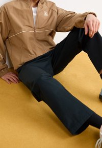 Brown zip-up jacket with white accents, worn over a white shirt, paired with black trousers. Sitting on a yellow floor.