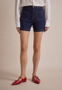Denim šorti ar augstu jostasvietu, tumši zila krāsa, dzeltenā šuvieniem un divām aizmugurējām kabatām, kopā ar sarkaniem zābakiem ar siksnu detaļu.