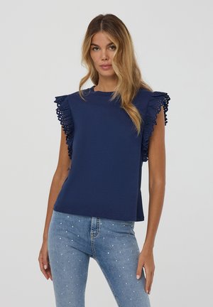 Top blu navy con dettagli in pizzo arricciato sulle spalle, dotato di scollatura rotonda e maniche corte, abbinato a jeans azzurri chiari decorati con borchie.