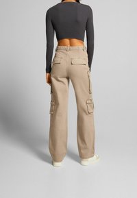 Pantalones cargo beige de cintura alta, con múltiples bolsillos y un corte relajado, combinados con una camiseta de manga larga ajustada de color oscuro.