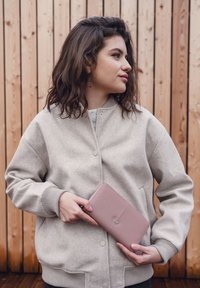 Light grey bomber jacket with a soft texture and ribbed cuffs. Holding a pale pink wallet with a silver accent, set against a wooden backdrop.