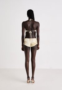 Gaiši beigu bikini tops ar saitēm kaklā un aizmugurē, kombinēts ar teksturētām zelta šortiem. Modelis valkā caurspīdīgas kurpes ar papēžiem.