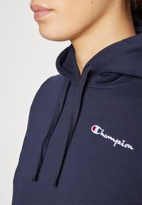 Hoodie blu navy con cordino, caratterizzato da un logo "Champion" ricamato in bianco e dettagli rossi, realizzato in morbido materiale misto cotone.