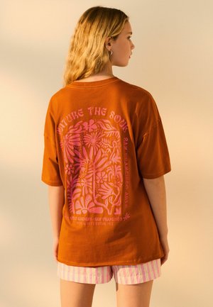 Brown oversized t-shirt featuring a colorful pink floral design on the back; paired with pink striped shorts. Soft cotton texture.