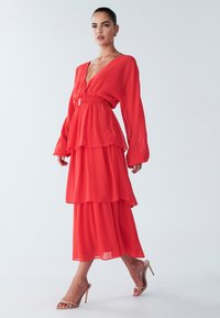 Abito in chiffon rosso a strati con maniche lunghe, scollatura a V e fascia elastica in vita. Presenta una silhouette fluente e morbida e delicati polsini sulle maniche.