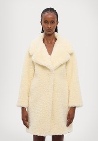 Cappotto teddy di colore crema, morbido e peloso, con un ampio colletto, maniche lunghe e una vestibilità ampia; presenta una texture soffice e senza chiusure visibili.