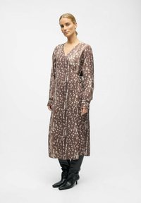 Femme aux cheveux blonds attachés, portant une robe midi à manches longues brun et blanc à pois, et des bottes noires pointues montantes jusqu'aux genoux.