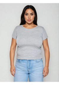 Kobieta o długich ciemnych włosach, ubraną w szarą koszulkę z krótkim rękawem i jasnoniebieskie dżinsy, stojąca na tle jednolitego jasnego tła.