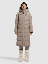 Lange beige gewatteerde jas met een capuchon, voorzien van een ritssluiting aan de voorkant, trekkoorden en doorgestikte secties. Gecombineerd met witte, geplooide sneeuwlaarzen.