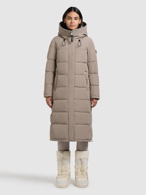 Długa beżowa pikowana kurtka puchowa z kapturem, zapinana na zamek, z detalami ze ściągaczami i przeszywanymi sekcjami. W komplecie białe, marszczone buty śniegowe.