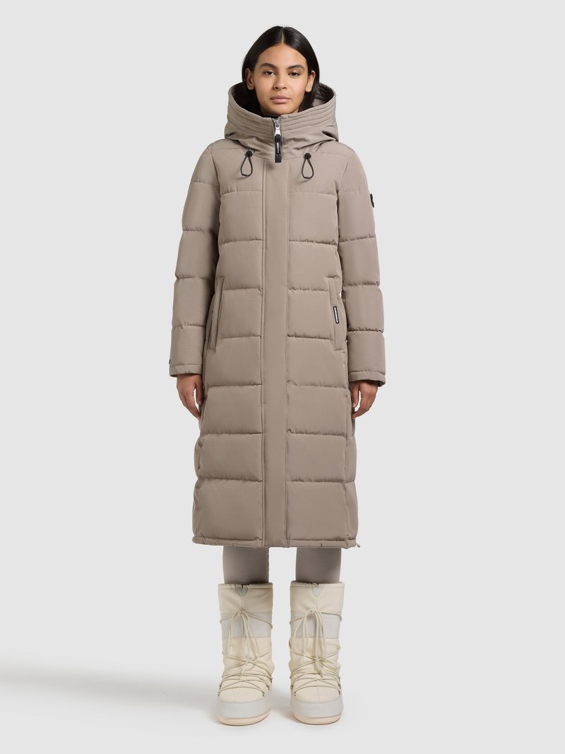 Lange beige gewatteerde jas met een capuchon, voorzien van een ritssluiting aan de voorkant, trekkoorden en doorgestikte secties. Gecombineerd met witte, geplooide sneeuwlaarzen.