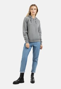 Grijze zip-up hoodie met trekkoorden, gestructureerde stof. Gecombineerd met lichtblauwe jeans met opgerolde manchetten en zwarte enkelboots, staande voor een witte achtergrond.