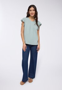 Blusa verde chiaro, fluida, con maniche a ruffle, abbinata a pantaloni in denim scuro a gamba larga. Modella che indossa sandali e anelli.