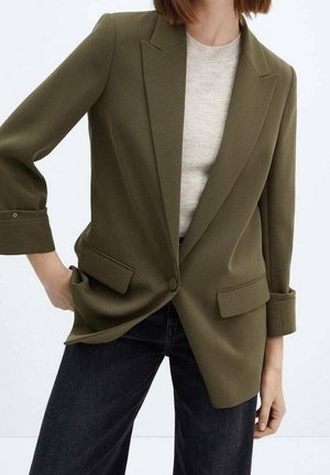 Kvinde iført en olivengrøn blazer over en lys beige sweater kombineret med mørke jeans, poserer med den ene hånd i lommen.