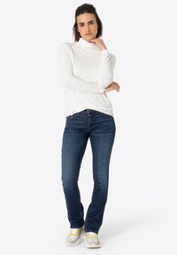 Witte turtleneck met lange mouwen, donkerblauwe jeans en sneakers met gele accenten. Gladde stof, getailleerd ontwerp, standaard denimsnit.