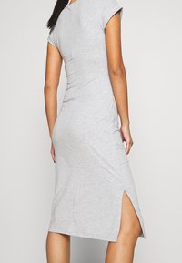 Femme portant une robe midi grise claire ajustée, avec des manches courtes et une fente haute sur le côté gauche, se tenant devant un fond uni.