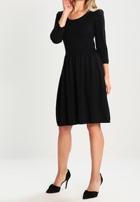 Donna che indossa un abito nero alla lunghezza del ginocchio con maniche tre quarti e scarpe col tacco alto nere, in piedi con una mano vicino al viso.