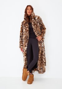 Manteau en fausse fourrure avec un motif léopard, doté d'un col montant. Leggings noirs et bottines beige complètent la tenue. Textures lisses présentes.