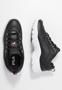Zapatillas deportivas negras con material de cuero liso, suela de goma blanca, ojales metálicos y detalle de logo en la lengüeta. Forma redondeada.