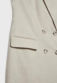 Blazer beige croisé avec quatre boutons en écaille et une poche à rabat sur le côté gauche.
