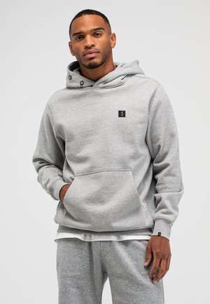 Mann trägt passenden hellgrauen Hoodie und Jogginghose, steht mit einer Hand in der Kapuzen-Tasche vor einem schlichten Hintergrund.