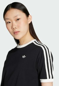 Black cotton t-shirt with white ribbed collar and three white stripes down the sleeves. Features a small embroidered logo on the chest.