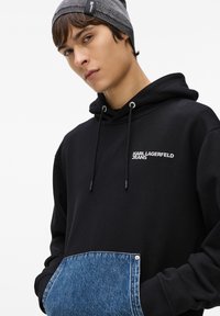 Juodas džemperis su pilku džinsiniu kišeniu, su raišteliais ir logotipu. Tekstūruotas pilkas skrybėlė papildyta apranga.