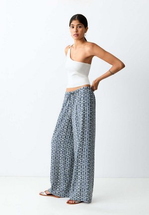 Mujer con un top blanco de un solo hombro, pantalones anchos azules con estampado y sandalias, de pie frente a un fondo blanco liso.