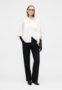 Λευκό φαρδύ πουκάμισο με τσέπη, συνδυασμένο με μαύρο παντελόνι. Το μοντέλο φοράει μαύρα δερμάτινα γάντια και μυτερά μαύρα παπούτσια, σε απλό φόντο.