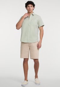 Camicia verde a quadri con maniche corte, modello con bottoni e tasca, abbinata a pantaloni corti in denim beige chiaro e sneakers bianche.