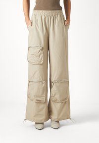 Beige Cargo-Hose mit weit geschnittenem Bein, elastischem Bund, zwei Fronttaschen und Reißverschlusstaschen an den Seiten. Hergestellt aus leichtem Stoff, mit verstellbaren Säumen.